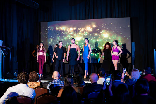 Models on a runway showcasing salon apparel at the grand opening of Stylist ProWear in Miami Lakes, with an audience watching and taking photos.