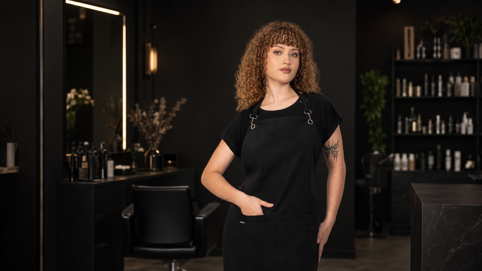 Model wearing a black salon apron in a modern dark salon with styling chairs, mirrors, and product shelves in the background.